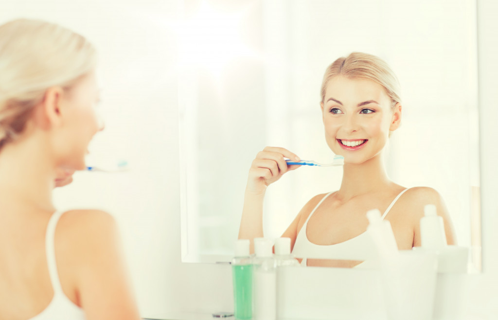  smiling young woman with toothbrush cleaning teeth and looking to mirror at home bathroom