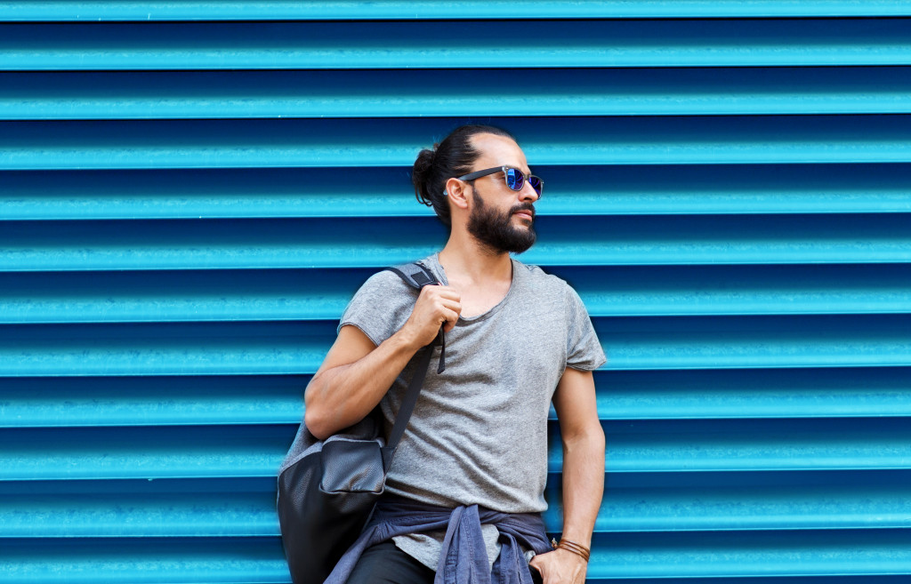 Young man with a beard and sunglasses holding a bag while standing against corrugated sheets.